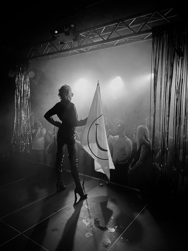 Schwarz weiss Bild zeigt die Berliner Drag Queen Amy Strong beim Auftritt in Drag Queen Show in Deutschland. Das Foto wurde von der Bühne hinter Amy in Richtung Publikum aufgenommen. Amy trägt eine blonde mittellange Perücke, eine große Sonnenbrille, ein enges, langärmliges Lederkleid und hochhackige Stiefel, die über das Knie reichen. In einer Hand hält sie eine weiße Fahne mit dem Smile-Logo. Amy steht aufrecht und schaut über das Publikum hinweg.